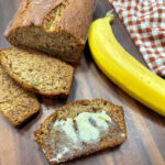 Banana bread on a cutting board with three slices. There's a banana next to the bread and a red and white gingham kitchen towel.