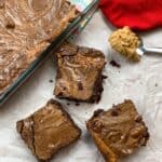 Three brownies sitting next to a pan of brownies and a spoonful of peanut butter