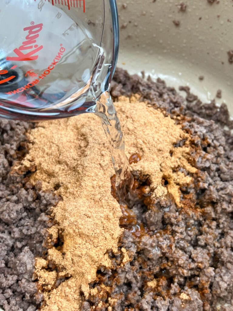 Pouring water into a saucepan with taco seasoning and ground beef. 