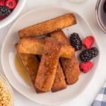 French toast sticks on a plate. There's a stick with maple syrup on it. The syrup is in a bowl. There are some berries next to the plate of French toast sticks.