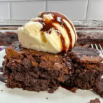 A brownie on a white plate with a scoop of ice cream. There's chocolate drizzled on top.