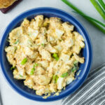 Egg salad in a blue bowl. There's a spoon on the side and some green onions on the other side.