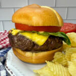 An air fryer burger on a bun with tomatoes and lettuce. There's some Ruffles chips on the white plate with the burger.