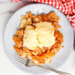 Apple dump cake on a white plate next to a red and white gingham towel. There's two scoops of ice cream right on top.