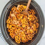 Chex Mix being stirred by a wooden spoon in a slow cooker.